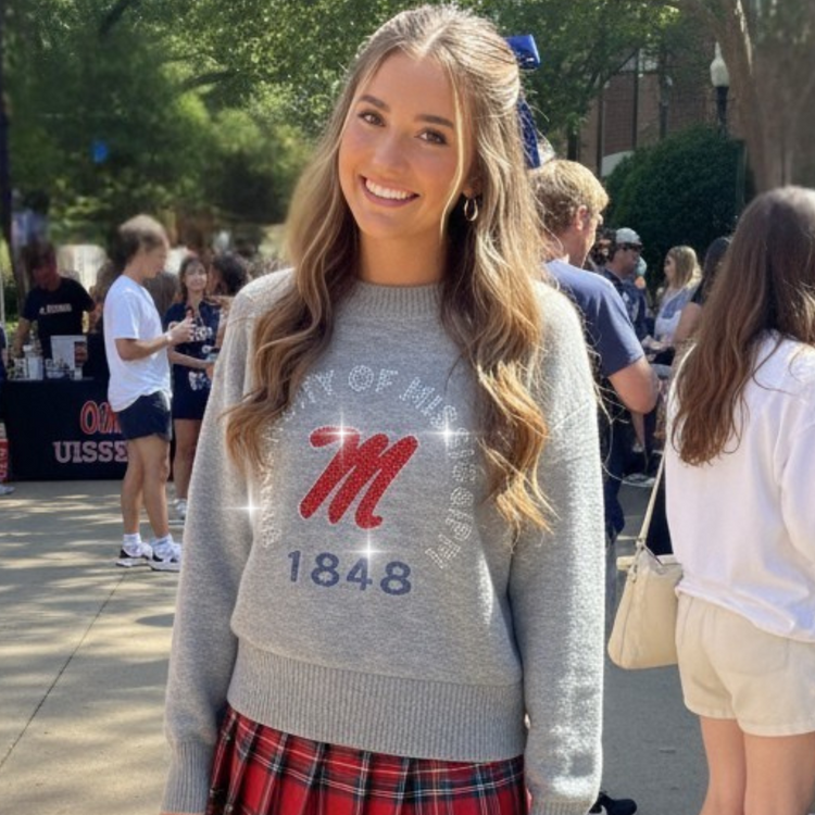 Woman wearing a gray sweater with a logo and plaid skirt in an outdoor setting