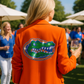 Person wearing an orange jacket with a gator logo, holding a glass of wine, in an outdoor setting.