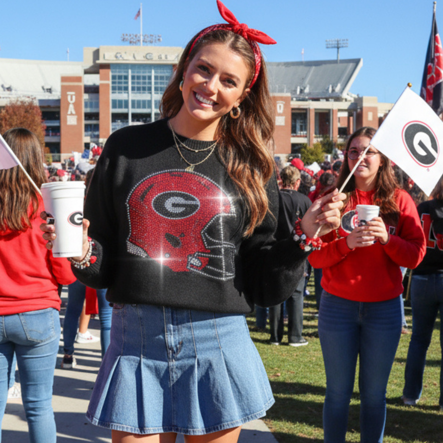 Woman at a sports event wearing a black sweater with a red logo and a denim skirt, holding a white cup.