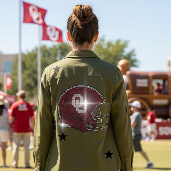 Person wearing a green jacket with a helmet design and stars, standing on a field with people and flags in the background.