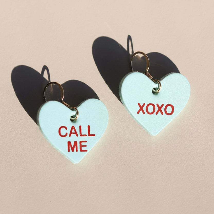 Heart-shaped earrings with 'Call Me' and 'XOXO' text on a beige background