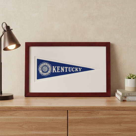 Framed Kentucky pennant on a wooden surface with a lamp and books in the background.