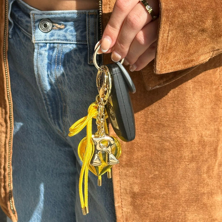 Keychain with yellow ribbon and gold charm held by a person wearing a brown coat and blue jeans.